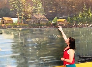 Kandi McGilton points to the fireweed in a painting of historic Metlakatla, painted by FM Harrow in 1930. The huge painting hangs on the back wall of the stage in Metlakatla's Town Hall. (Photo by Leila Kheiry.) 