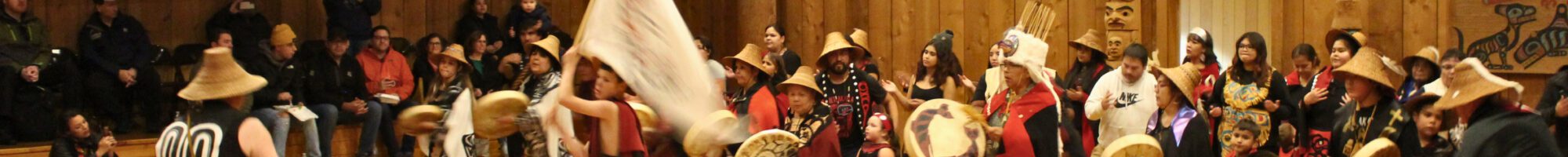 a group of people in traditional red and black regalia dance, sing and play drums in a large, open, wooden building